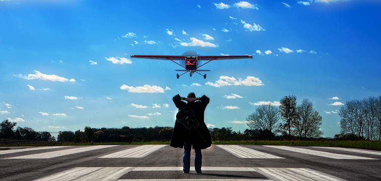 David Sloan standing on airport runway as plane comes in, and of course, with guitar