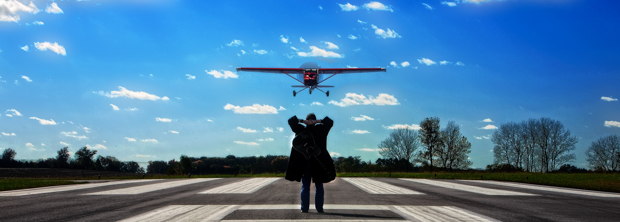 David Sloan standing on airport runway as plane comes in, and of course, with guitar